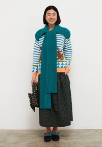 Hrubá tyrkysová šatka prehodená cez pruhovaný sveter s hnedo-zelenými vzormi, doplnená čiernou kockovanou sukňou a tyrkysovými topánkami.