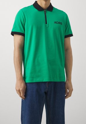 Mężczyzna w zielonej koszulce polo z krótkim rękawem, czarnym kołnierzykiem i mankietami, czarnym zamkiem błyskawicznym, napisem "KORS" na piersi oraz w niebieskich dżinsach.