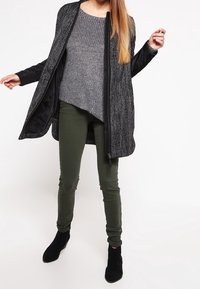 Donna che indossa un cappotto grigio texture sopra un maglione grigio a maglia, pantaloni verde oliva e stivaletti neri, in piedi contro uno sfondo bianco.