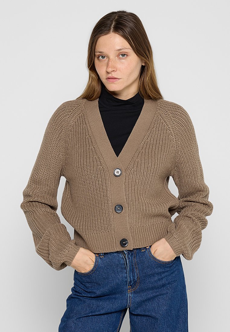 Brauner gestrickter Cardigan mit tiefem V-Ausschnitt, geripptem Muster, langen Ärmeln und drei dunklen Knöpfen vorne. Kombiniert mit hochtaillierten Jeans.