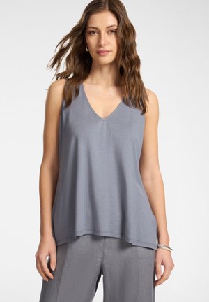 Kobieta z falującymi brązowymi włosami, ubrana w luźny szary bezrękawnik z dekoltem w serek i dopasowane szare spodnie, stojąca na jednolitym tle.