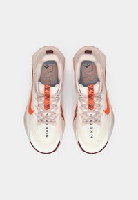 Nike running shoes in light tan with orange accents. Mesh upper, textured pattern on the sole, and visible branding on the tongue and side.