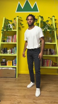 T-shirt blanc, jean noir, et baskets blanches. L'arrière-plan présente un mur jaune, des étagères vertes avec des livres et des plantes.