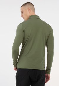 Uomo con capelli corti che indossa una camicia a maniche lunghe verde oliva e pantaloni neri, di spalle rispetto a uno sfondo bianco uniforme.