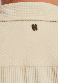 Beige strukturiertes Gewebe mit einem geriffelten Muster. Es verfügt über ein schwarzes Etikett mit der Gravur "PME LEGEND", das mit einem Faden befestigt ist.