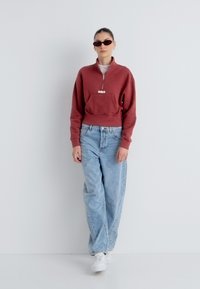 Κόκκινο cropped φούτερ με φερμουάρ μπροστά και μπροστινή τσέπη, σε συνδυασμό με ανοιχτό μπλε φαρδιά τζιν και λευκά αθλητικά παπούτσια. Το μοντέλο φοράει μαύρα γυαλιά ηλίου.