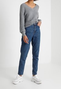 Personne portant un pull en maille gris à col en V, un jean taille haute bleu et des baskets blanches épaisses, debout avec une main dans la poche.