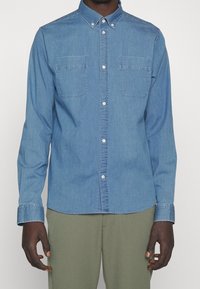 Camisa de denim azul claro con mangas largas, cuello tipo camisero, dos bolsillos en el pecho y botones blancos en contraste. Textura suave y corte ajustado.