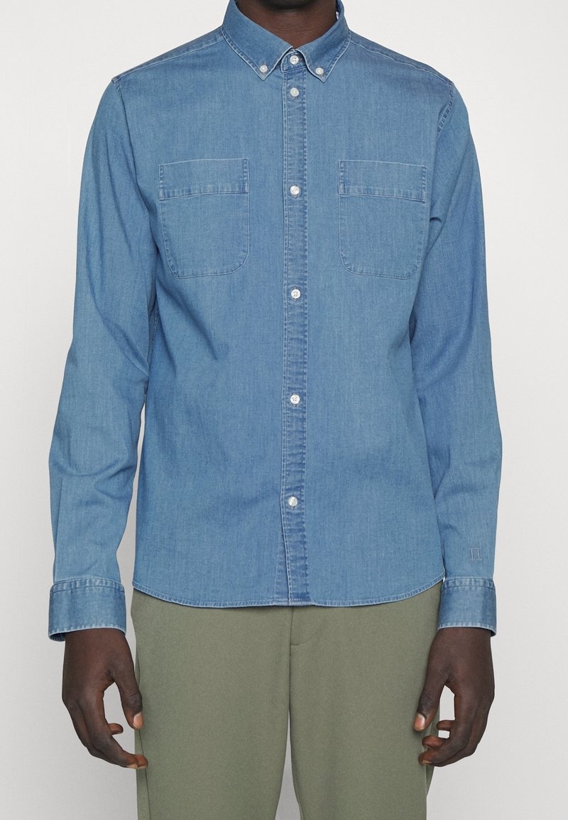 Camisa de denim azul claro con mangas largas, cuello tipo camisero, dos bolsillos en el pecho y botones blancos en contraste. Textura suave y corte ajustado.