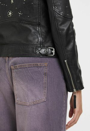 Schwarze Lederjacke mit silbernen Sternverzierungen, strukturierter Oberfläche und Reißverschlussärmeln. Dazu hellviolette Jeans mit Muster.