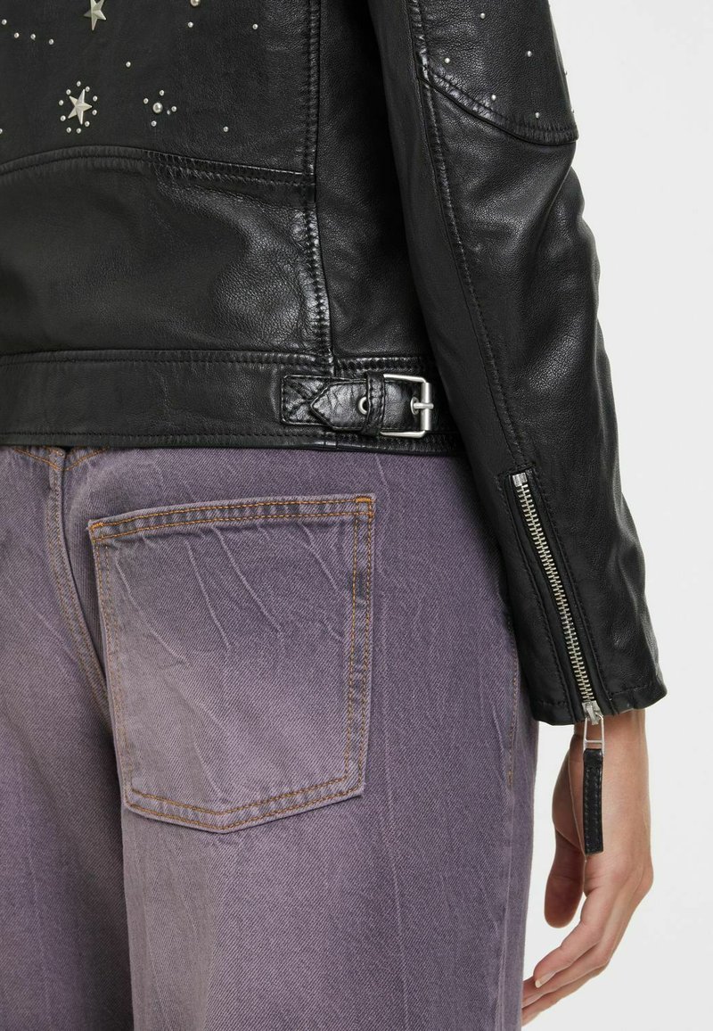 Black leather jacket featuring silver star embellishments, textured finish, and zippered cuffs. Paired with light purple patterned jeans.