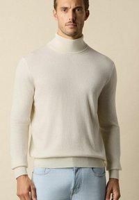 Sweter w kolorze kremowym z golfem, wykonany z miękkiej, fakturowanej dzianiny, z ribbowymi mankietami, w połączeniu z jasnoniebieskimi dżinsami. Prosty i nowoczesny design.
