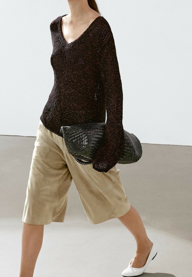 Personne portant un pull en maille marron foncé ample et un short beige mi-long, portant un sac tissé noir, marchant en chaussures plates blanches.