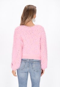 Puszysty różowy sweter z okrągłym dekoltem, luźnymi rękawami i prążkowanym dołem, z wielokolorowymi plamkami dla dodatkowej tekstury. Noszony z niebieskimi dżinsami.