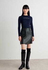 Tengerészkék hosszúujjú póló "Tommy Jeans" logóval, fekete bőr mini szoknyával, fekete térd feletti zoknival és fekete magassarkú csizmával párosítva.