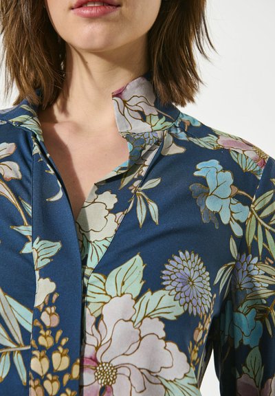 Femme portant un chemisier bleu foncé avec un motif floral coloré, montrant le col et la partie supérieure de la poitrine, avec des cheveux courts et bruns.