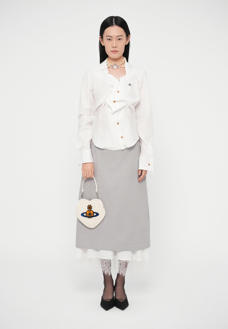 Mujer con camisa blanca abotonada, falda gris sobre leggings blancos de encaje, zapatos negros, collar de perlas y sosteniendo un bolso blanco en forma de corazón.