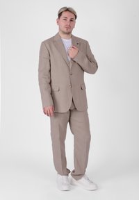 Completo in lino beige chiaro con due bottoni, pantaloni abbinati e una t-shirt bianca semplice. Calzature: sneaker bianche. Sfondo semplice.