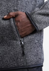 Giacca grigia con motivo a spina di pesce e tasca con zip; presenta una rifinitura nera liscia e un tessuto resistente per il calore.