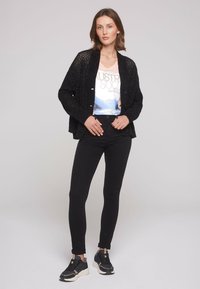Un cardigan noir en tricot avec des détails de boutons porté sur un t-shirt graphique, associé à un jean skinny noir et des baskets noires. Texture lisse, design décontracté.
