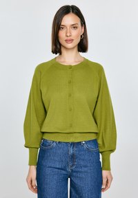 Žena s rovnými hnědými vlasy nosí olivově zelený zapnutý cardigan s nafouklými rukávy a modré džíny s vysokým pasem, stojící před bílým pozadím.