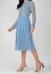 Vestido azul claro de longitud hasta la rodilla con un cuerpo de perlas y mangas largas transparentes. La falda es fluida con una textura fruncida. Combinado con tacones metálicos.