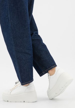 Persona che indossa jeans blu scuro e sneakers bianche con suola piatta che fa un passo avanti su uno sfondo bianco semplice.