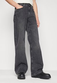 Jean en denim noir à jambes larges avec un délavage, comportant des poches avant et une coupe décontractée, associés à des bottes noires épaisses.