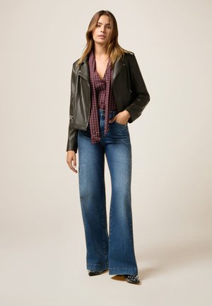 Veste en cuir noir sur une chemise à carreaux, assortie à un jean en denim à jambes larges décoré de clous. La tenue est complétée par des chaussures pointues.