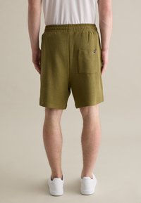 Grüne, strukturierte Shorts mit elastischem Bund und einer Gesäßtasche, kombiniert mit weißen Sneakers, von hinten gezeigt.