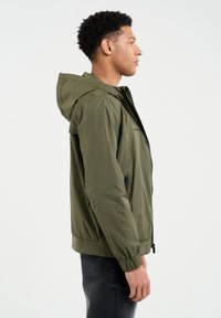 Olivinio žalia sukapota striukė su lygiu, lengvu tekstūru. Yra elastingi rankogaliai ir apačia, juodos detalės bei užtrauktuko priekinis uždarymas.