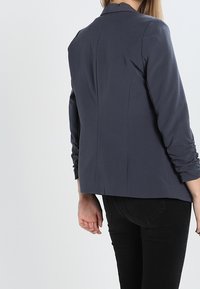 Mörkgrå blazer med en figursydd design, slagkrage och rynkade ärmar. Slät tyg med en skräddarsydd bakdel och inga synliga knäppningar.