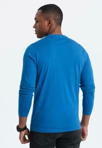 Camicia blu a maniche lunghe realizzata in tessuto morbido, con collo a giro e design aderente. Il retro presenta una texture liscia con dettagli minimi.