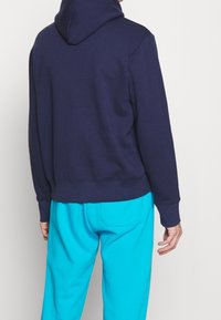 Mörkblå hoodie med dragsko i huvan, ribbade muddar och fåll. Bärs med ljus turkosa byxor som visar en avslappnad passform och mjuk textur.