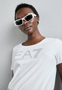 T-shirt bianco in cotone con logo grigio EAT Emporio Armani in strass; abbinato a occhiali da sole rettangolari bianchi e orecchini d'oro.