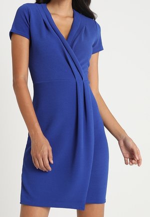 Kobieta ubrana w krótką, sięgającą kolan sukienkę kopertową w kolorze królewskim niebieskim, z dekoltem w serek i plisowanym przodem.
