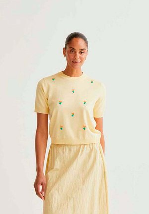 Mujer con el cabello recogido, vestida con un top amarillo pálido con pequeños bordados florales y una falda amarilla a juego, de pie frente a un fondo claro.