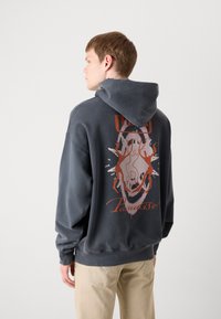 Sudadera con capucha gris que presenta una impresión gráfica de un rostro y formas abstractas en naranja y blanco. Tejido suave, corte relajado.