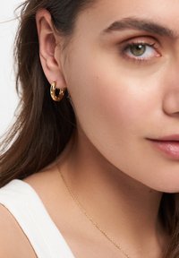 Goldene Creolen zeichnen sich durch ein elegantes, glänzendes Design mit einer glatten Oberfläche aus. Das Modell hat lange Haare, und eine zarte Goldkette ist sichtbar.