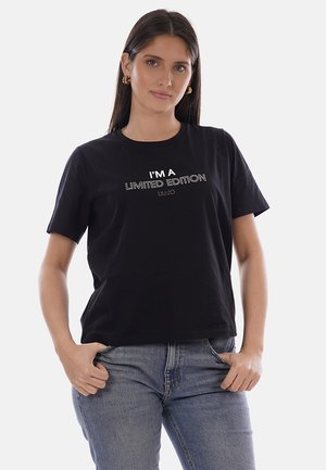 Donna con lunghi capelli scuri, che indossa orecchini a cerchio dorati, una maglietta nera con la scritta "SONO UN'EDIZIONE LIMITATA" e jeans blu, con le mani nelle tasche.