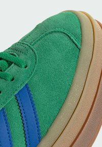 Sapatilhas de camurça verdes com detalhes em pele azul, uma entressola texturizada e sola de borracha de goma contrastante com formato arredondado.