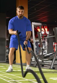 Corde da battaglia nere tenute da una persona in maglietta blu e pantaloni corti, mentre si allena su una superficie in erba verde con attrezzature da palestra sullo sfondo.