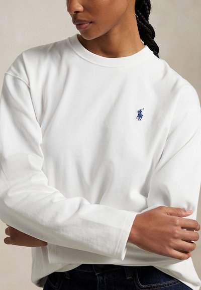 Osoba z warkoczami nosi białą koszulę z długim rękawem z małym niebieskim logo gracza polo na piersi, z rękami skrzyżowanymi z przodu.