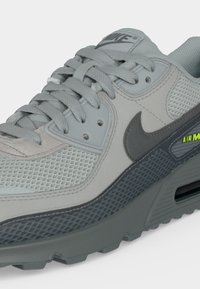 Γκρι αθλητικό παπούτσι Nike Air Max με επάνω μέρος από δίχτυ, σκούρο γκρι σχέδιο Swoosh, υφασμένη ενδιάμεση σόλα και κορδόνια δεμένα πάνω από τη μαξιλαρωμένη γλώσσα.