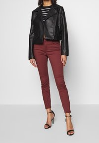 Chaqueta de moto de cuero negra corta, camiseta a rayas negra y blanca, pantalones ajustados burdeos y tacones de aguja negros con banda en el tobillo.