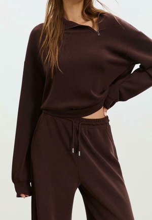 Persoon draagt een donkergroene top met lange mouwen en ritskraag en bijpassende broek met trekkoord, hand op de heup, met los haar over de schouder.