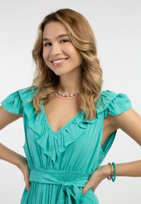 Rochie turcoaz cu volane, decolteu adânc în V, talie strânsă și colier și brățări colorate, asortate. Material neted, accente vibrante.