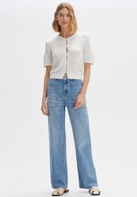 Weiß strukturierte, kurzärmelige Strickjacke mit Knopfdetails, kombiniert mit hellblauen, weit geschnittenen Jeans und schwarz-weißen Rutsch-Sandalen.