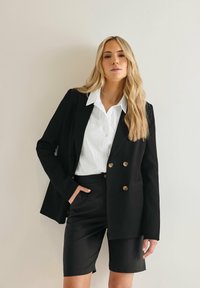 Svart kavaj med guld dubbla knappar över en vit skjorta med krage, kombinerad med svarta skräddarsydda shorts. Slitstarkt tyg, modern passform.