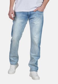 Man som bär ljusblå urtvättade jeans och vita sneakers, med en hand i fickan och vit skjorta.